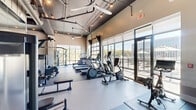 Located at 4810 Bradford Dr NW in Huntsville, Alabama, this modern fitness center offers a generously sized, single-floor exercise space designed to support a comprehensive range of workout routines. The facility features an industrial-inspired aesthetic with exposed ductwork, concrete structural elements, and dark-painted ceilings that lend the space a contemporary, loft-like character. Large floor-to-ceiling windows line the perimeter, flooding the interior with natural light and offering views of the surrounding outdoor greenery and adjacent outdoor patio area.

The exercise room is divided into distinct functional zones. The cardio area is equipped with multiple treadmills, elliptical machines, and a stationary bike arranged along the windowed walls. A dedicated strength training zone features a multi-station cable machine, a power rack with TRX suspension trainers, a heavy punching bag, and a functional trainer. The free weights section includes a full dumbbell rack and adjustable benches, complemented by a medicine ball and kettlebell storage station. Floor-to-ceiling mirrors expand the sense of space and assist with form monitoring. Athletic flooring throughout provides durability and comfort. Motivational large-format wall murals add an energetic visual element, completing the dynamic and well-appointed atmosphere of this fitness facility.