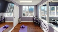 The Mills Crossing Townhomes & Apartments fitness center is a thoughtfully designed, single-floor amenity space that offers residents a well-rounded workout experience. The interior is finished with distinctive lavender-painted walls, warm hardwood-style flooring, and a drop-panel ceiling, creating a clean and energizing environment throughout.

The space is divided into two functional areas. The yoga and stretching studio features a large wall-mounted mirror, a wall-mounted television for guided workout sessions, and ample open floor space accommodating yoga mats and foam blocks. Natural light streams in through multiple windows, offering pleasant views of the surrounding neighborhood and filling the room with a bright, motivating ambiance.

Adjacent to the studio, a dedicated weight and cardio room houses a multi-station cable machine, treadmills, and additional fitness equipment. A pass-through window connects the two areas, enhancing the open and airy feel of the overall space.

The entrance is welcoming, featuring a branded Mills Crossing entry mat at the glass-paneled door, flanked by sidelights that further maximize natural light. The fitness center is well-equipped and efficiently laid out, providing residents with a convenient and comfortable space to support a healthy, active lifestyle.