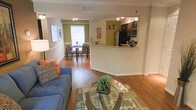 This single-floor residence showcases an open-concept layout that effortlessly connects the living, dining, and kitchen areas into one cohesive and functional space. Rich hardwood flooring flows throughout, anchoring the warm and welcoming ambiance. The living area is bright and comfortable, accented by crown molding and a classic chair rail detail that adds architectural character to the walls.

The kitchen features dark cabinetry contrasted by light granite countertops and a breakfast bar that serves as a natural dividing element between the cooking and living spaces. Stainless steel appliances, including a refrigerator and microwave, add a modern touch, while track lighting overhead ensures excellent task illumination.

Adjacent to the living space, the dining area benefits from natural light streaming through a large window, creating an airy atmosphere ideal for everyday meals or entertaining guests. An accent wall in a warm olive green tone adds a pop of color and visual depth to the space.

Overall, the layout is thoughtfully designed to maximize flow and connectivity, making the space feel expansive and easy to navigate. The combination of classic architectural details, modern finishes, and a cohesive color palette gives this residence a balanced, timeless appeal suited for comfortable everyday living.