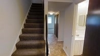 This two-story residence offers a straightforward and functional layout distributed across two floors. The interior is accessed through a central hallway that connects the main living areas, finished with tile flooring in a neutral, earthy tone that provides durability and easy maintenance. A carpeted staircase with a dark handrail leads to the upper level, offering a contrast between the soft textile of the stairs and the hard flooring below.

The hallway extends toward what appears to be a carpeted room at the far end, visible through a doorway fitted with a sliding door — likely a bedroom or utility space. A ceiling-mounted light fixture provides overhead illumination in the corridor, supplemented by natural light filtering in through a window at the end of the hall.

To the right of the staircase, a partial view of a bathroom is visible, featuring white cabinetry and a sink, suggesting a well-appointed and conveniently located bathroom on the main floor. The walls throughout are painted in a soft, neutral gray-blue tone, contributing to a calm and cohesive interior aesthetic. Overall, the space presents a practical and livable environment across its two floors.