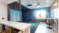 This single-floor indoor golf simulator space is thoughtfully designed to deliver an immersive and functional golfing experience. The primary simulator bay is enclosed by dark, floor-to-ceiling curtains that create an ideal projection environment, with a large high-definition screen displaying realistic course visuals. Two ceiling-mounted projectors and a launch monitor system are positioned overhead to capture and analyze each shot with precision. A synthetic turf hitting mat sits atop dark carpet flooring, providing a realistic feel underfoot, while multiple golf bags with clubs are neatly stored beside a natural-light window that offers views of the surrounding landscape. The adjacent area transitions seamlessly into a brighter, more utilitarian space finished with warm light wood flooring, recessed lighting, and clean white walls. This area features a tall storage cabinet, a wall-mounted workstation, and access to additional rooms, including what appears to be a sauna or wellness space visible through a glazed door. Framed artwork and paneled wood cabinetry add a polished, contemporary aesthetic throughout. A thermostat and occupancy signage near the entrance indicate the space is designed for shared or commercial use. Overall, the facility balances high-performance sports technology with a clean, modern interior design.