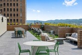 This rooftop amenity space combines a spacious interior with a generous open-air terrace, delivering an elevated urban experience with sweeping panoramic views of the surrounding cityscape and beyond. The interior is designed with a clean, modern aesthetic, featuring light hardwood flooring, white walls, and recessed lighting that create a bright, welcoming atmosphere throughout.

The open-plan interior is divided into distinct functional zones. A fully equipped kitchenette runs along one wall, featuring warm wood cabinetry, a sleek white quartz island with bar seating, stainless steel appliances including a full-size refrigerator, an undermount sink with a matte black faucet, and a dishwasher. Adjacent to the kitchen, a generous dining and gathering area accommodates multiple seating configurations, including communal tables and casual café-style arrangements. A comfortable lounge zone offers blue modular sofas and accent seating, while a separate media room provides a wall-mounted television, plush sectional sofas, and direct terrace access via floor-to-ceiling sliding glass doors.

The outdoor terrace is the defining feature of this space, extending the usable area considerably. Finished with large-format pavers and framed by warm wood privacy planters, it is furnished with a variety of café tables and lounge seating. Floor-to-ceiling glass walls and oversized sliding doors seamlessly connect the interior to the terrace, flooding the space with natural light and unobstructed city views.