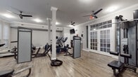 This fitness center is thoughtfully designed to support a comprehensive workout experience. The space features light hardwood-style flooring throughout, complemented by neutral gray walls and crisp white crown molding that give the room a clean, polished appearance. Decorative white columns with detailed capitals add an architectural character rarely found in fitness facilities.

The cardio section is well-appointed with treadmills, elliptical trainers, and a stationary bike, providing ample options for aerobic exercise. The strength training area includes multiple cable and resistance machines, a bench press station, and functional training accessories such as battle ropes, catering to a wide range of fitness routines. A full wall of mirrors along one side of the room enhances the sense of openness and allows users to monitor their form during workouts.

Natural light fills portions of the space through large bay windows and glass-paned French doors, creating a bright and inviting environment. Multiple ceiling fans are installed throughout, ensuring comfortable airflow. A wall-mounted television is positioned for easy viewing during cardio sessions. Built-in shelving provides storage for accessories, and the overall layout allows for generous circulation between equipment stations, making this fitness center both functional and comfortable to use.