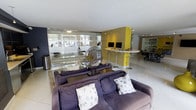 This single-floor space showcases a thoughtfully designed open-plan layout that seamlessly blends comfort and contemporary style. The living area is anchored by plush purple sofas adorned with decorative patterned cushions, complemented by a glass coffee table that adds an airy, modern touch. A striking sculptural centerpiece sits prominently on the coffee table, serving as a bold artistic focal point.

A vibrant yellow accent wall draws the eye toward a built-in workstation area equipped with dual monitors and a sleek bar-height white countertop with transparent acrylic bar stools — ideal for casual work or collaboration. This area is illuminated by recessed track lighting that highlights the space's clean lines and modern aesthetic.

Large floor-to-ceiling glass partitions allow natural light to filter through while offering views of the surrounding exterior greenery, creating a bright and open atmosphere throughout. The white tile flooring runs consistently across the space, reinforcing a clean, cohesive look. A secondary lounge area is visible in the background, further demonstrating the generous, flowing layout. Overall, the space exudes a polished, functional, and design-forward character.