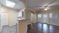 This single-story unit offers a well-organized layout connecting the kitchen, living area, bedroom, and bathroom in a cohesive flow. The open living room is a standout space, featuring rich dark hardwood-style flooring, crown molding, and a ceiling fan. Natural light fills the room through a large window and a set of French doors that provide direct outdoor views and access. Track lighting along one wall adds supplemental illumination.

The kitchen is positioned adjacent to the living area and defined by a curved peninsula with granite-style countertops, providing a natural division between spaces. White cabinetry lines the walls, complemented by a white electric range, built-in microwave, and dishwasher. A skylight above the kitchen area introduces additional natural light.

The bedroom is carpeted and benefits from crown molding, a ceiling fan, and a window that invites natural light. The bathroom connects directly to a walk-in closet and features granite-style countertops, a single-basin vanity, white cabinetry, and a bathtub. The same dark hardwood-style flooring extends through the bathroom, maintaining a consistent aesthetic throughout the unit.