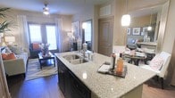 This single-floor residence showcases a thoughtfully designed open-concept layout that seamlessly connects the kitchen, dining, and living areas. The kitchen serves as the centerpiece, anchored by a substantial island topped with light granite countertops and fitted with a deep double-basin stainless steel sink. Dark cabinetry provides a striking contrast against the stone surfaces, while elegant pendant lighting illuminates the workspace from above. The open flow leads directly into a warm and inviting living area, where rich hardwood flooring extends throughout, complemented by neutral, earthy wall tones that create a cohesive and welcoming ambiance. A ceiling fan overhead adds both function and a finishing touch to the living space. Natural light fills the interior through multiple windows dressed with blinds and sheer curtains, offering a bright and airy atmosphere. A door visible from the central corridor suggests well-defined private spaces beyond the main living area. The overall design balances modern finishes with a comfortable, livable aesthetic, making it a practical yet refined space suited for everyday living and entertaining alike.