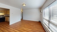 This single-floor unit presents a clean, straightforward layout with consistent design elements carried throughout its rooms. The living area is generously proportioned, featuring warm-toned hardwood-style flooring that extends throughout the space, creating a cohesive and inviting atmosphere. Large windows dressed with horizontal blinds allow natural light to fill the room while offering views of the surrounding greenery. A wall-mounted air conditioning unit and baseboard heating system provide year-round climate control, and a flush-mount ceiling light fixture provides overhead illumination.

The bedroom maintains the same hardwood-style flooring and white walls, with a window that frames outdoor tree views, contributing to a calm and airy feel. Baseboard heating is present here as well, ensuring consistent comfort.

The kitchen is equipped with light-toned cabinetry offering ample storage above and below the countertops, which feature a dark laminate surface. A gas range with an overhead ventilation hood serves as the focal point, and the layout provides practical counter space on both sides for food preparation. The flooring continues seamlessly from the main living areas.

The bathroom includes a full-size soaking tub with an overhead shower and a curtain rod, surrounded by clean white square tile that lines the walls of the tub enclosure, giving the space a classic, utilitarian appearance.