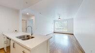 This single-floor residence is designed with a clean, modern aesthetic, characterized by crisp white walls, warm hardwood-style flooring, and an abundance of natural light throughout. The open-concept living and kitchen area forms the heart of the space, creating a seamless flow between both zones. The living area is generously proportioned, featuring a ceiling fan with integrated lighting and a large floor-to-ceiling window dressed with vertical blinds, offering views of surrounding greenery while filling the space with natural light.

The kitchen is efficiently laid out with a peninsula island that doubles as a prep and casual dining space. It features white quartz-style countertops, a double stainless steel sink with a contemporary pull-down faucet, white lower cabinetry, warm wood-toned upper cabinets, and a full suite of stainless steel appliances including an electric range, over-the-range microwave, and refrigerator.

The bedroom is a well-proportioned, carpeted room with a large window that mirrors the natural light found throughout the unit. The bathroom is clean and functional, featuring hardwood-style flooring, a soaking tub with an overhead shower, and simple, bright white finishes that lend a fresh, airy feel to the room.