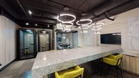 Located at 26 E Chestnut St in Chicago's vibrant Near North Side, this contemporary coworking lounge delivers a thoughtfully designed, single-floor workspace that balances collaboration and privacy. The space makes a strong first impression with its industrial-chic aesthetic — exposed dark ceiling beams and ductwork contrast with clean white walls and warm, textured slatted wood accent panels throughout.

At the heart of the lounge sits a commanding communal work surface finished in dramatic veined stone, surrounded by bold yellow accent chairs that inject energy into the space. Striking circular ring pendant lights hang overhead, creating a dynamic focal point. A wall-mounted display and built-in cabinetry with stone countertops provide additional functionality along the perimeter.

Fully enclosed glass-walled conference rooms offer private meeting settings while maintaining visual connectivity to the broader space. Compact acoustic pods provide quiet individual retreats for focused work or calls. A comfortable lounge area with soft seating and a dedicated wet bar with stone backsplash and custom cabinetry rounds out the amenities.

Patterned carpet tiles, layered lighting, and sculptural white wall art add depth and sophistication throughout. The overall ambiance is professional yet inviting, making this an ideal setting for productivity and collaboration in the heart of Chicago.
