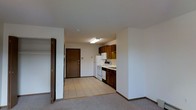 This single-floor unit offers a functional and straightforward layout designed for comfortable everyday living. The open-concept design connects the carpeted living area to the kitchen, creating a cohesive and practical space. The kitchen is equipped with dark wood cabinetry that provides generous storage, complemented by white appliances including a refrigerator, electric range, and dishwasher. A flush overhead light fixture brightens the kitchen area, ensuring a well-lit workspace. The transition between the living area and kitchen is marked by a shift from plush carpet to durable tile flooring, offering both comfort and easy maintenance where it matters most. A solid wood entry door leads into the unit, with a light-colored wall panel nearby, likely housing a thermostat or intercom. Adjacent to the living area, a bifold closet door opens to reveal a built-in storage space complete with a hanging rod, ideal for organization. The neutral white walls and light-toned surfaces throughout contribute to a clean, understated ambiance that is easy to personalize. Overall, the space is designed with practicality in mind, making efficient use of every area.