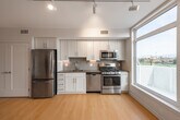 This contemporary single-floor residence showcases a clean, modern design throughout, unified by warm light wood flooring and crisp white walls that create a bright, airy atmosphere. The well-appointed kitchen is a standout feature, offering white shaker-style cabinetry extending to the ceiling, dark countertops, a tiled backsplash, and a full suite of stainless steel appliances including a gas range, built-in microwave, dishwasher, and refrigerator. Large windows in the kitchen and living areas flood the space with natural light and frame open views of a green outdoor landscape, including athletic fields and distant scenery. The living area is generously sized, featuring track lighting and direct access to a private balcony through glass doors, extending the living space outdoors. A separate room with a closet and an electrical panel provides additional utility and storage. The bathroom is finished to a high standard, featuring a spacious glass-enclosed walk-in shower clad in large-format gray tile with a built-in niche and wall-mounted lighting, reflecting a polished, contemporary aesthetic. Overall, the space delivers a cohesive modern living environment with quality finishes and an abundance of natural light.