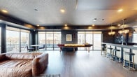 This sophisticated amenity lounge is designed to offer an elevated social experience, combining refined aesthetics with versatile entertainment features. The space is anchored by a striking wet bar area, where a substantial stone-topped counter is lined with sleek gold-framed barstools beneath pendant lighting and a patterned gold ceiling that adds warmth and visual interest. Mirrored cabinetry behind the bar extends the sense of depth while reinforcing the upscale atmosphere. Adjacent to the bar, a dedicated prep and service area features dark cabinetry with ample countertop workspace, framed by city views through large windows.

The lounge area is defined by wide-plank hardwood flooring, deep charcoal walls, and a geometrically patterned ceiling that lends architectural texture throughout. A full-size shuffleboard table serves as a central focal point, complemented by a leather sofa, accent chairs, and a media wall with a large flat-screen television. Floor-to-ceiling sliding glass doors line the perimeter, flooding the interior with natural light and connecting seamlessly to a spacious outdoor terrace furnished with dining and lounge seating. The terrace offers panoramic urban skyline views, making it an ideal setting for both casual gatherings and private events.