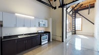 This 2-bedroom, 1-bathroom loft showcases a striking industrial aesthetic rooted in converted warehouse architecture. The open living area commands attention with dramatically soaring ceilings supported by exposed black steel trusses and warm wood roof decking, creating a grand, airy atmosphere throughout. Polished concrete floors extend seamlessly across the main living space, reflecting light and reinforcing the loft's clean, contemporary character.

The kitchen is thoughtfully appointed with two-tone cabinetry — crisp light grey upper cabinets contrasting with deep espresso lower cabinets — paired with dark granite countertops and a full suite of stainless steel appliances, including a gas range, over-the-range microwave, and refrigerator. Original architectural details, including white-painted exposed brick columns and bold black steel structural elements, add industrial character throughout.

The expansive main living area offers generous open space, with a staircase providing access to upper-level areas. The bathroom features a dark granite vanity counter, a white under-mount sink, white cabinetry, and polished flooring, maintaining the consistent modern aesthetic. Large industrial-style windows along the perimeter allow natural light to fill the space, enhancing the loft's open, urban feel.