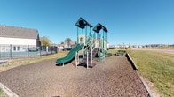 Located at 815 Cole Dr in Johnstown, Ohio, this community outdoor space centers around a well-equipped playground designed to serve the surrounding residential neighborhood. The play area features a prominent dual-tower structure finished in green and tan, complete with two slides, staircase access, and elevated platforms topped with pitched roof canopies. The structure provides a variety of play opportunities for children of different ages and abilities.

The playground surface is covered with rubber mulch, providing a safety-conscious ground covering beneath and around the play equipment. A park bench is positioned nearby, offering a comfortable seating area for caregivers and visitors to observe the play space. The area is bordered by a black metal fence along one side, clearly defining the space and providing an added layer of safety.

The surrounding landscape is open and grassy, offering room to roam beyond the core play structure. The neighborhood's residential homes are visible in the background, reinforcing the community-oriented character of the space. Under clear open skies, the playground benefits from ample natural light throughout the day, making it an inviting and accessible outdoor destination for families in the Johnstown area.