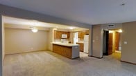 This single-floor space offers a comfortable and functional open-concept layout that seamlessly connects the living and kitchen areas. The generously sized living room is finished with soft wall-to-wall carpeting and neutral gray walls, creating a warm and welcoming atmosphere. A classic chandelier adds a touch of elegance to the space, complementing the recessed lighting throughout.

The kitchen is thoughtfully designed with warm honey-toned oak cabinetry providing ample storage, paired with light countertops that offer plenty of prep space. A peninsula-style breakfast bar extends from the kitchen, creating a natural divide between the cooking and living areas while also serving as a convenient spot for casual dining or entertaining. The kitchen is equipped with white appliances, including a built-in microwave, a range, and a full-sized refrigerator. Tile flooring in the kitchen provides a practical contrast to the carpeted living area.

A hallway visible from the main living space leads to additional rooms, suggesting a well-organized floor plan. The open sightlines throughout the space and the combination of recessed and pendant lighting ensure a bright, airy feel. Overall, the space presents a clean, classic interior that balances practicality with everyday comfort.