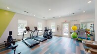 Located at 5000 Seaforth St in Shallotte, NC, this fitness center offers a thoughtfully designed workout environment within a compact, efficiently utilized single-floor footprint. The space features crisp white walls accented by a bold yellow geometric wall mural, adding an energetic visual element that complements the active atmosphere.

The flooring throughout is a striking multi-toned vinyl plank design in blue, tan, and warm neutral tones, providing both durability and a modern aesthetic. Recessed lighting illuminates the space evenly, while multiple windows along the walls invite natural light and outdoor views, creating an open and inviting feel.

Cardio equipment includes multiple treadmills and an elliptical trainer, all from reputable brands. A rowing machine rounds out the cardio offerings. Strength and flexibility training is supported by a cable/weight machine, a dumbbell rack with bench, and an assortment of stability balls in vibrant yellow and green. A large wall mirror expands the sense of space and aids in form monitoring during workouts.

Practical amenities include a dual-level water fountain and bottle-filling station mounted to the wall, ensuring easy hydration access. A glass-paneled door provides a convenient direct exit to the exterior. The overall design is clean, bright, and functional, making this fitness center a well-appointed resource for residents.