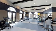 The West Ranch Fitness Center in Carlsbad is a thoughtfully designed, single-floor facility built to support a comprehensive workout experience. The space features soaring ceilings accented by dark exposed wood beams, which contrast elegantly against the white ceiling and neutral-toned walls, creating a refined yet energizing atmosphere. Large arched windows with dark frames line the walls, allowing an abundance of natural light to flood the interior while offering open views of the surrounding landscape. French doors provide additional access points and contribute to the bright, airy feel of the space. The flooring consists of durable, two-toned carpet tiles in shades of grey and blue, designed to accommodate high-traffic fitness use. A ceiling fan is centrally mounted to ensure comfortable air circulation throughout. The facility is well-stocked with a range of cardio equipment, including multiple treadmills and rowing machines, as well as strength training stations and a medicine ball rack. Recessed lighting supplements the natural light, ensuring the space remains well-lit at all hours. The overall ambiance is clean, spacious, and welcoming, making it a fully functional and visually appealing fitness destination for residents of the West Ranch community.