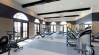 The West Ranch Fitness Center in Carlsbad is a thoughtfully designed, single-floor facility built to support a comprehensive workout experience. The space features soaring ceilings accented by dark exposed wood beams, which contrast elegantly against the white ceiling and neutral-toned walls, creating a refined yet energizing atmosphere. Large arched windows with dark frames line the walls, allowing an abundance of natural light to flood the interior while offering open views of the surrounding landscape. French doors provide additional access points and contribute to the bright, airy feel of the space. The flooring consists of durable, two-toned carpet tiles in shades of grey and blue, designed to accommodate high-traffic fitness use. A ceiling fan is centrally mounted to ensure comfortable air circulation throughout. The facility is well-stocked with a range of cardio equipment, including multiple treadmills and rowing machines, as well as strength training stations and a medicine ball rack. Recessed lighting supplements the natural light, ensuring the space remains well-lit at all hours. The overall ambiance is clean, spacious, and welcoming, making it a fully functional and visually appealing fitness destination for residents of the West Ranch community.
