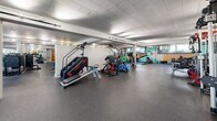 Located at 13900 Tahiti Way in Marina del Rey, California, this fitness facility spans a single floor and is organized into two dedicated exercise rooms, together offering an impressively comprehensive workout environment. The main exercise room is a spacious, open-plan area with durable rubber flooring, structural columns, and a tiled drop ceiling with track and recessed lighting that ensures consistent, even illumination throughout. Windows along the perimeter admit natural light and views of the surrounding landscape. The room is equipped with a wide array of strength-training machines, a full dumbbell rack, weight benches, barbells with bumper plates, cable systems, a stair climber, treadmills, stationary bikes, and additional cardio equipment. Large wall mirrors extend visibility across the floor, enhancing the sense of space. Vibrant geometric murals in teal, orange, and black punctuate the walls, adding an energetic visual character to the environment. Built-in cubbies provide personal storage for users. The second exercise room is dedicated to functional and combat fitness, featuring suspended heavy punching bags, gymnastics rings, exercise mats, stability balls, and plyometric boxes, all within a clean, mirror-lined space with bright overhead lighting. Together, the two rooms form a well-rounded, full-service fitness facility suited for a wide range of training disciplines.