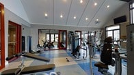 This fitness center offers a comprehensive workout environment designed to accommodate a wide range of exercise needs. The space is outfitted with an array of strength training machines, including cable and pulley systems, as well as cardio equipment such as stationary bikes, providing members with versatile options for their fitness routines. The room features large mirrors framed in red along one wall, which serve both a functional and aesthetic purpose — enhancing the sense of space while allowing users to monitor their form during exercise. The distinctive multi-colored floor, composed of blue, tan, and white geometric sections, adds a dynamic visual energy to the space. Overhead, recessed globe lighting is arranged across a paneled ceiling, providing bright, even illumination throughout. Crown molding along the upper walls adds a refined architectural touch to the otherwise utilitarian environment. The facility is single-level and benefits from natural light filtering in through windows visible in the mirrored reflection, contributing to a welcoming and energizing atmosphere. Overall, the fitness center strikes a balance between functional equipment layout and clean, purposeful design.