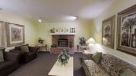 This single-floor space offers a welcoming and comfortable atmosphere centered around a traditional brick fireplace, which serves as the room's focal point. The fireplace features a clean white mantel adorned with framed artwork, candlesticks, and decorative accents, adding a refined touch to the rustic brick surround. Soft yellow-green walls contribute to the room's warm and calming ambiance, complemented by recessed track lighting overhead and elegant crown molding that frames the ceiling. The seating area is furnished with a patterned sofa accented by green throw pillows, alongside a dark upholstered chair, creating an inviting arrangement around a white rectangular coffee table. Fresh floral arrangements in soft blues and pinks are thoughtfully placed throughout, enhancing the room's welcoming character. Two side tables flanking the fireplace hold additional floral décor and a classic table lamp, providing gentle ambient lighting. A framed landscape artwork on the wall adds a finishing artistic element. The plush dark carpeting underfoot ties the space together, lending a sense of warmth and comfort throughout.