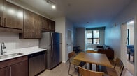Located at 135 E Grand River Ave in East Lansing, MI, this single-floor unit offers a modern, urban design with consistent finishes throughout. The kitchen is well-appointed with dark espresso cabinetry extending to the ceiling, white quartz countertops, and a full suite of stainless steel appliances including a refrigerator, dishwasher, electric range, and over-the-range microwave. Light wood-tone flooring runs throughout the main living areas, complementing the contemporary aesthetic. The kitchen opens directly into a dining area and living room, creating a cohesive, functional layout. The living room features exposed ductwork details and large windows that bring in natural light and outdoor views. The bedroom is a private, enclosed space with a large window offering additional natural light. Two bathrooms serve the unit: one features a glass-enclosed walk-in shower with wood-grain tile surround, and the other includes a dual-sink vanity — both finished with dark cabinetry, white countertops, and light wood-tone flooring. Track lighting in the kitchen and clean neutral wall tones throughout contribute to the space's modern, understated character.