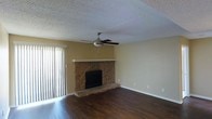 This single-floor space centers around a striking stone fireplace that serves as the focal point of the main living area. The fireplace features a natural stacked-stone surround with a raised hearth and a clean stone mantel, adding rustic character and warmth to the room. Rich, dark hardwood flooring runs throughout, providing a polished and durable foundation that contrasts nicely with the warm beige tones of the walls. A ceiling fan with a brushed nickel finish is centrally mounted, offering both air circulation and a subtle lighting element. The textured popcorn ceiling adds a classic finishing detail overhead. Natural light enters the space through a set of sliding glass doors fitted with vertical blinds, brightening the room and providing potential access to an outdoor area. The open layout creates a sense of spaciousness, making the living area feel both comfortable and versatile. Neutral wall colors and clean baseboards contribute to the overall understated, move-in-ready aesthetic of the space.