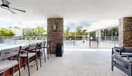 Located at 22555 SW 107th Ave in Goulds, Florida, this single-story community amenity facility offers a well-appointed outdoor and indoor experience designed for leisure and social gathering. At the heart of the space is a expansive resort-style swimming pool surrounded by a generous concrete deck, lounge chairs, a shaded umbrella seating area, and lush tropical landscaping including mature palm trees and manicured hedges that create a private, resort-like atmosphere. The pool area is secured by a sleek metal perimeter fence with gated access. An adjoining covered pavilion provides a shaded outdoor lounge space furnished with contemporary modular seating and a bar-height counter area, anchored by a striking stone-clad column with wall-mounted lighting. Large floor-to-ceiling sliding glass windows and doors connect the covered pavilion to the interior, filling both spaces with natural light and maintaining visual continuity between indoors and out. The interior features clean white walls, polished concrete flooring, built-in cabinetry with warm wood accents, and ceiling fans throughout. A water fountain station and additional seating areas are integrated along the corridor. The overall design aesthetic is clean, modern, and functional, reflecting a South Florida lifestyle focused on outdoor living.