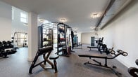 Located at Heritage Towne Blvd in Midlothian, TX, this two-story fitness center offers a spacious and well-appointed workout facility designed to accommodate a wide range of exercise needs. The main floor is generously sized, featuring soaring double-height ceilings in key areas, floor-to-ceiling windows that flood the space with natural light and provide views of the surrounding outdoors, and durable rubber flooring throughout. This level is comprehensively equipped with Matrix-brand strength machines, a multi-functional cable and free-weight rig, benches, barbells, weight racks, and an assortment of cardio equipment including treadmills, ellipticals, and an air bike. Circular pendant lighting adds a modern aesthetic touch. A dedicated area on the main floor also houses a water fountain station for convenience. The upper floor, accessed via an open staircase, offers a more intimate exercise space with additional cardio equipment including rowing machines and stationary bikes, exposed ductwork lending an industrial character, and a railing overlooking the main floor below. Both floors maintain a clean, modern design with white walls and neutral tones, creating a bright and motivating environment for fitness pursuits.