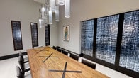 This single-floor space presents a refined and professional environment centered around a standout conference room. The room is anchored by a substantial wood conference table showcasing rich grain detail and distinctive X-shaped metal inlay accents, surrounded by leather upholstered chairs that seat a generous number of attendees. Overhead, a dramatic cluster of cylindrical glass pendant lights cascades from the ceiling, creating a layered, gallery-like lighting effect that elevates the room's ambiance. The neutral gray walls serve as a sophisticated backdrop for carefully curated artwork, including a framed figurative print and a series of bold, textured abstract panels with intricate cross-hatched patterns in deep blue and black tones, lending the space a contemporary artistic character. The light-toned flooring complements the overall modern aesthetic, while dark-framed accent panels add visual depth and contrast throughout. The high ceiling contributes to an open, airy feel despite the room's intimate and focused function. Together, the design elements create a space that balances creativity with professionalism, making it well-suited for meetings, presentations, or collaborative gatherings that benefit from an inspiring and polished environment.