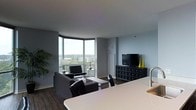 This single-floor space showcases a contemporary open-concept design that integrates the kitchen and living areas into one cohesive environment. The living room is anchored by a striking corner window arrangement that spans nearly floor to ceiling, flooding the space with natural light and offering expansive outdoor views of the surrounding landscape and skyline. The grey-toned walls complement the dark hardwood flooring, creating a sleek, modern aesthetic throughout.

The living area is furnished with a tufted black leather seating arrangement, centered around a glass-top coffee table, and accompanied by a classic Barcelona-style accent chair. A substantial dark media console with multi-drawer storage houses a flat-screen television and is accented by tall yellow decorative vases, adding a bold pop of color to the otherwise neutral palette.

The kitchen is visible in the foreground, featuring a white countertop island and a modern brushed-metal faucet, contributing to the clean, minimalist design. The open layout between the kitchen and living room enhances the sense of spaciousness and flow. Overall, the space exudes a polished, urban aesthetic with thoughtful design details throughout.