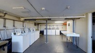 Located at 99 Hemingway Ave in East Haven, Connecticut, this shared laundry facility offers a practical and functional space designed to serve building residents efficiently. The single-floor room features a generous layout with clearly defined zones for washing, drying, and folding. A row of top-load washing machines lines one wall, while a separate bank of dryers sits opposite, providing ample capacity for simultaneous use. Utility sinks are conveniently available along the back wall, offering additional washing options for hand-wash items or general cleaning tasks. A large folding table with metal hairpin legs provides a dedicated workspace for sorting and folding laundry. The concrete floor is durable and easy to maintain, suited to the demands of a high-traffic utility space. Overhead fluorescent lighting ensures the room is well-lit throughout. Exposed pipes and ductwork run along the ceiling, contributing to the utilitarian character of the space. Support columns are positioned throughout the room without obstructing workflow. Safety is addressed with a visible security camera mounted on the ceiling. Informational signage is posted on the walls, keeping users informed of facility guidelines. Overall, the space is straightforward, well-equipped, and organized for efficient daily use.