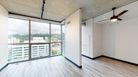 Located at 333 SW Harrison St in Portland, Oregon, this single-floor unit showcases a contemporary industrial-modern design characterized by exposed concrete ceilings and light wood-style flooring throughout. The open layout is immediately defined by expansive floor-to-ceiling glass walls that frame panoramic views of Portland's cityscape and the surrounding tree-covered hills, allowing abundant natural light to fill the living areas.

The main living space is open and versatile, with partial partition walls creating defined zones without sacrificing the airy, connected feel of the interior. Track lighting mounted to the concrete ceiling provides flexible illumination across the living and sleeping areas. The kitchen is efficiently designed with white upper cabinetry, teal-accented lower cabinetry, stainless steel appliances including a refrigerator and microwave, and clean white countertops. An entry corridor connects to the main living areas and includes access to interior doors.

The bathroom features a full soaking tub with shower combination, clean white surround panels, and chrome fixtures. The unit's consistent use of light-toned flooring, crisp white walls, and raw concrete ceilings creates a cohesive, modern aesthetic with an industrial sensibility suited to urban living.