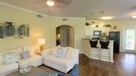 This single-floor residence offers an open-concept layout that integrates the living, dining, and kitchen areas into one cohesive space. The living room is anchored by rich hardwood flooring, a ceiling fan, and crown molding details that add a refined touch to the warm, neutral-toned walls. A distinctive arched doorway leads to an adjacent room, likely a study or den, visible in the background and adding architectural character to the space.

The kitchen is positioned at the rear of the open floor plan and features white cabinetry, a dark granite or stone countertop, and stainless steel and black appliances. Pendant lighting hangs above the counter, illuminating a breakfast bar fitted with sleek, modern bar stools—an ideal casual dining spot.

Natural and artificial lighting work together to create a bright, inviting ambiance throughout. The overall design blends classic architectural elements, such as the arched entryway and crown molding, with contemporary finishes, resulting in a balanced and functional living environment suited for everyday comfort and entertaining.