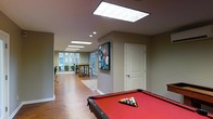 This single-floor amenity space is thoughtfully designed to accommodate both recreation and casual gatherings. The layout flows seamlessly from one zone to the next, beginning with a dedicated billiards area anchored by a full-size pool table with a striking red felt surface. Hardwood-style flooring runs throughout the space, lending warmth and cohesion to the entire area. The walls are finished in a neutral taupe tone, providing a clean, modern backdrop that highlights the colorful accents, including a vibrant billiard-themed artwork that serves as a focal point. Overhead, large recessed panel light fixtures and skylights flood the interior with bright, even illumination, creating a welcoming atmosphere at any time of day. Natural light also enters through generous windows at the far end of the space, where a casual dining or meeting nook features a wooden table and chairs set atop a patterned area rug. A private door along one wall suggests access to an additional room or utility space. The open-plan design encourages a social environment, making this space well-suited for community recreation, events, or everyday leisure activities.