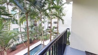 This single-floor space includes a private balcony that offers a standout feature: a picturesque view of a beautifully landscaped tropical courtyard. The balcony is fitted with a sleek, dark metal railing that provides both safety and a modern aesthetic contrast to the natural surroundings. From this vantage point, towering palm trees rise prominently, their slender trunks and lush canopies creating a verdant, resort-like atmosphere. Below, a narrow water feature winds through the courtyard, adding a tranquil, reflective element to the outdoor scenery. The landscaping is well-maintained, with layered tropical plantings and reddish mulch ground cover completing the lush setting. The surrounding building exterior is finished in a clean, light stucco, with additional balconies visible across the courtyard, suggesting a thoughtfully designed community environment. The overall ambiance is calm and tropical, with abundant greenery providing a natural sense of privacy and seclusion. The balcony itself benefits from open-air exposure and natural light filtered through the palm canopy, making it an inviting outdoor extension of the living space.