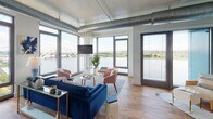Located at 1900 Half St SW in Washington, DC's vibrant Southwest Waterfront neighborhood, this modern residence showcases an industrial-chic aesthetic defined by exposed silver ductwork, soaring ceilings, and warm hardwood flooring throughout. The expansive living area is the centerpiece of the unit, wrapped in floor-to-ceiling windows that frame breathtaking panoramic views of the Potomac River and the iconic Frederick Douglass Memorial Bridge, flooding the space with abundant natural light. The open-concept kitchen is outfitted with white cabinetry, dark stone countertops, a full suite of stainless steel appliances, and track lighting overhead. The bedrooms continue the industrial design language with exposed ductwork overhead and large windows — one offering sweeping waterfront views, another overlooking the dynamic surrounding cityscape — complemented by hardwood floors and generous closet access. Two well-appointed bathrooms feature white cabinetry with dark countertops, penny-tile flooring, and clean white tile work, with one offering a soaking tub and shower combination and the other a glass-enclosed walk-in shower. The overall ambiance is contemporary and sophisticated, blending urban energy with serene waterfront tranquility.