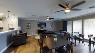 This single-floor space offers an open-concept layout that seamlessly combines living and dining areas into one cohesive, functional environment. The interior is finished with warm hardwood-style flooring that runs throughout, complementing the cool gray walls and crisp white crown molding that frames the ceiling with a clean, polished look.

The living area is anchored by a large, dark leather sectional sofa arranged around a wall-mounted flat-screen television, which sits atop a sleek media console. Botanical wall art in framed panels adds a touch of nature-inspired décor to the space. Two ceiling fans with integrated lighting fixtures are evenly distributed across the room, ensuring both comfort and adequate illumination.

The dining area features two dark-toned dining tables accompanied by wooden chairs, providing ample seating for gatherings. Natural light filters into the space through windows dressed with horizontal blinds and dark curtain panels, offering both brightness and privacy control.

Recessed lighting in the ceiling supplements the ceiling fans, creating a layered lighting scheme. The overall aesthetic is contemporary and understated, combining dark furnishings with neutral wall tones to create a sophisticated yet comfortable atmosphere well-suited for both everyday use and entertaining.