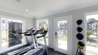 This single-floor fitness center is designed to provide a clean, functional, and inviting workout environment. The space is finished with neutral gray walls and crisp white crown molding and door trim, lending it a modern, polished aesthetic. Recessed lighting in the smooth white ceiling ensures consistent illumination throughout the room, complementing the generous natural light that pours in through multiple large windows. The windows frame pleasant views of the surrounding outdoor area, including greenery and neighboring structures, creating an open and airy atmosphere that enhances the workout experience. A glass-paneled door with a secure electronic lock provides direct access to an outdoor recreational space, seamlessly connecting the interior to the exterior. The fitness area is equipped with multiple treadmills featuring built-in digital consoles, positioned to take advantage of the natural light and outdoor views. The flooring is finished in a dark wood-look material, providing a durable and stylish surface suited for active use. Overall, the space is thoughtfully laid out to maximize both functionality and comfort, making it a practical and welcoming facility for daily exercise.