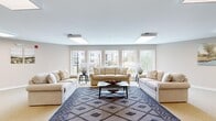 Located at 6101 Barboursville Lane in North Chesterfield, Virginia, this single-floor community room offers a well-appointed, multi-functional space designed for gatherings and everyday use. The open layout is organized into distinct zones that flow seamlessly into one another, creating a comfortable and practical environment.

The lounge area is anchored by a set of neutral-toned sofas arranged around a patterned area rug and a central coffee table. Large windows along the perimeter flood this zone with natural light and provide outdoor views of the surrounding community. Flush-mounted panel lighting supplements the natural light throughout.

Transitioning toward the interior, multiple round dining tables with modern black chairs provide ample seating for group meals or meetings. A support column subtly defines the boundary between the lounge and dining areas without interrupting the open feel of the space. A separate dining nook, framed by interior windows overlooking an adjacent room, offers a more intimate setting.

The kitchenette is outfitted with warm-toned wood cabinetry, a double stainless steel sink, a microwave, and a full-size black refrigerator, making the space well-equipped for hosting events. Neutral carpet flooring and light beige walls contribute to the room's calm, welcoming ambiance throughout.