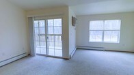 This single-floor residential space offers a clean, functional layout centered around a spacious, light-filled living area. The room is finished with wall-to-wall carpeting that adds warmth underfoot, while neutral-toned walls provide a versatile backdrop suitable for a variety of interior styles. A prominent set of sliding glass doors serves as the focal point of the main living area, opening directly onto a wooden deck that overlooks a peaceful, wooded landscape — ideal for enjoying natural scenery in every season. Adjacent to the sliding doors, a generously sized window on the opposite wall further enhances the room's bright and airy atmosphere by inviting additional natural light throughout the day. A wall-mounted HVAC unit and baseboard heating are both visible, ensuring year-round climate comfort. The open layout of the living area provides flexible arrangement options, making the space easily adaptable to different furniture configurations and lifestyle needs. The overall ambiance is calm and understated, with the surrounding tree line visible through multiple windows creating a tranquil, nature-connected feel throughout the interior.