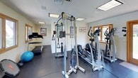 This dedicated fitness facility is thoughtfully designed to support a full range of workout routines. The space features durable rubber mat flooring throughout, providing a safe and cushioned surface for exercise. Bright flush-panel skylights and recessed lighting ensure the room is well-lit at all times, while multiple wood-framed windows and a set of French doors invite natural light and offer views of the surrounding outdoor greenery and pool area.

The strength training section is comprehensively outfitted with a multi-station cable machine, a squat/power rack, a full dumbbell rack ranging across multiple weight increments, and an adjustable weight bench. Cardio equipment includes a treadmill and an elliptical trainer, catering to a variety of aerobic preferences. Stability and functional training tools such as a stability ball, BOSU ball, and TRX-style suspension straps are also on hand.

A large wall-mounted mirror enhances the sense of space and allows for form monitoring during workouts. A wall-mounted flat-screen television provides entertainment options. An adjacent room, accessible through a glass-paneled wood door, appears to serve as a supplementary lounge or equipment area. The overall ambiance is clean, functional, and welcoming, making this a versatile space for fitness enthusiasts of all levels.