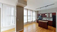 This single-floor residence is defined by its open, airy layout and consistent design choices throughout. Warm honey-toned hardwood flooring runs continuously across the living and kitchen areas, creating a cohesive and inviting foundation. Large floor-to-ceiling windows fitted with vertical blinds line the main living space, flooding the interior with abundant natural light and reinforcing the sense of openness.

The kitchen is a functional and well-appointed space, showcasing rich cherry wood cabinetry with both frosted and solid panel upper doors, complemented by a mosaic tile backsplash in neutral tones. Black appliances, including a range, built-in microwave, dishwasher, and refrigerator, provide a bold contrast to the lighter countertops. A freestanding island with a granite-style surface offers additional prep space and casual dining potential. Pendant lights hang from a track lighting system overhead, providing targeted illumination.

The living area is open and flexible, with a structural column adding architectural interest while defining distinct zones within the space. The bathroom features a full soaking tub with an overhead shower, white subway tile surround, and chrome fixtures, maintaining the clean, modern aesthetic found throughout the unit.