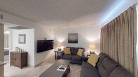 Located in Scottsdale, Arizona, this single-floor unit offers a thoughtfully arranged layout that connects the living, dining, kitchen, and sleeping areas cohesively. The living room serves as the central gathering space, furnished with two charcoal-gray sofas accented by mustard yellow throw pillows, a wall-mounted flat-screen TV, and warm ambient lighting from flanking table lamps. Light tile flooring runs throughout, contributing to a clean, airy feel, while a textured ceiling adds a classic Arizona residential touch.

Adjacent to the living room, the dining area features a dark wood table with seating for four and a raised bar-height counter with stools that connect directly to the kitchen. The kitchen is efficiently designed with white shaker-style cabinetry, granite-look laminate countertops, a stainless steel sink, and white appliances including a range and microwave, making it fully functional for everyday use.

The bedroom is a calm retreat, anchored by a queen-sized bed with a white upholstered headboard, framed artwork above, wall-mounted sconce lighting, and a ceiling fan. A wall-mounted TV and built-in dresser add practicality. The adjoining bathroom features a wide vanity with an extended mirror, white cabinetry, and tile flooring, maintaining the unit's consistent clean aesthetic throughout.