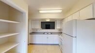 This two-floor space offers a straightforward, functional layout with consistent finishes throughout. The kitchen is equipped with white cabinetry extending to the ceiling, gray laminate countertops, a double-basin sink, a gas range, dishwasher, and a full-size refrigerator. Light tile flooring runs underfoot, and a pass-through window connects the kitchen to the adjacent living area, enhancing the sense of openness between the two spaces. The living area features blue-gray carpeting, neutral walls, and a window that brings in natural light with views of surrounding greenery. A distinctive green entry door adds a subtle accent to the otherwise neutral palette. Carpeted rooms throughout the upper level maintain the same understated aesthetic, with windows providing additional natural light and outdoor views. The bathroom includes a tiled floor with a classic blue accent dot pattern, a full tub and shower combination, and a natural light source via a window. A separate vanity area features a double-sink setup with a wide mirror and ample cabinet storage below. A visible electrical panel is accessible near the kitchen area. Textured ceilings are present throughout the space.