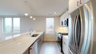 Located at 396 Cambridge St in Boston's Allston neighborhood, this single-floor unit showcases a modern, open-concept design finished with light hardwood flooring and neutral tones throughout. The layout is efficiently organized, connecting the living area, kitchen, and bedrooms in a cohesive flow.

The galley-style kitchen is well-appointed with stainless steel appliances, including a gas range, over-the-range microwave, and French door refrigerator. White shaker cabinetry reaches toward the ceiling, complemented by a white subway tile backsplash and light quartz countertops. Pendant lighting and recessed fixtures provide layered illumination, while a window above the sink introduces natural light and city views.

The open living area is bright and spacious, featuring recessed lighting and large windows alongside a glass-paned door that leads to a private balcony with views of the surrounding Boston streetscape. A stacked washer and dryer unit is conveniently tucked into a dedicated closet off the main living space.

The bedrooms are generously sized with light hardwood floors and ample closet space. The bathroom features large-format dark floor tiles, white subway tile surrounding the soaking tub and shower combination, and a clean white vanity, delivering a crisp, contemporary aesthetic throughout.