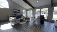 This single-story space showcases a thoughtfully designed interior that blends contemporary style with functional versatility. The vaulted ceiling, accented by a striking exposed dark wood beam, adds architectural character and a sense of openness throughout the main area. Durable hardwood-style flooring in a rich grey tone runs throughout, providing a sleek, cohesive foundation.

The space is divided into two distinct functional zones. A comfortable lounge area features modern upholstered seating arranged around a round ottoman atop a circular area rug, complemented by a wall-mounted flat-screen television and a built-in beverage station with a wine cooler and countertop appliances along one wall.

Adjacent to the lounge, a workspace area includes a dark executive desk with accompanying guest seating, ideal for meetings or consultations. Large floor-to-ceiling windows and a glass door flood the interior with natural light while offering direct views of an outdoor pool area and surrounding landscaping. The neutral grey color palette, clean lines, and modern furnishings throughout contribute to a polished, professional ambiance.
