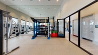 This contemporary fitness center is thoughtfully designed to accommodate a wide range of workout styles and fitness goals. The space features soaring ceilings that create an open, airy atmosphere, while large windows along the walls flood the interior with natural light and offer views of the surrounding area. Durable speckled rubber flooring runs throughout, providing a safe and resilient surface for all types of exercise.

At the heart of the gym stands a striking blue functional training rig, fully loaded with a dumbbell rack, battle ropes, resistance bands, medicine balls, and TRX suspension trainers — ideal for functional and high-intensity training. Colorful plyo boxes add to the versatility of the space. Floor-to-ceiling mirrors line the walls, visually expanding the room and allowing users to monitor their form.

The strength training area is equipped with a cable machine, barbell racks, weight benches, and a selection of free weights. The cardio zone features multiple treadmills, ellipticals, a stationary bike, and a stair climber, all positioned to take advantage of the natural light and outdoor views. A wall-mounted television and bold blue accent wall with framed artwork add a polished, motivating aesthetic. A glass-panel door provides a seamless connection to an adjacent area, completing this comprehensive and inviting fitness facility.