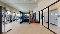 This contemporary fitness center is thoughtfully designed to accommodate a wide range of workout styles and fitness goals. The space features soaring ceilings that create an open, airy atmosphere, while large windows along the walls flood the interior with natural light and offer views of the surrounding area. Durable speckled rubber flooring runs throughout, providing a safe and resilient surface for all types of exercise.

At the heart of the gym stands a striking blue functional training rig, fully loaded with a dumbbell rack, battle ropes, resistance bands, medicine balls, and TRX suspension trainers — ideal for functional and high-intensity training. Colorful plyo boxes add to the versatility of the space. Floor-to-ceiling mirrors line the walls, visually expanding the room and allowing users to monitor their form.

The strength training area is equipped with a cable machine, barbell racks, weight benches, and a selection of free weights. The cardio zone features multiple treadmills, ellipticals, a stationary bike, and a stair climber, all positioned to take advantage of the natural light and outdoor views. A wall-mounted television and bold blue accent wall with framed artwork add a polished, motivating aesthetic. A glass-panel door provides a seamless connection to an adjacent area, completing this comprehensive and inviting fitness facility.