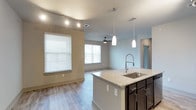 This single-floor space showcases a thoughtfully designed open-concept layout where the kitchen and living area flow seamlessly together. The kitchen is anchored by a central island topped with light granite countertops and equipped with a stainless steel undermount sink and a professional-style pull-down faucet. Dark espresso cabinetry beneath the island provides a striking contrast to the light countertop surfaces. Elegant pendant lights hang above the island, complementing the track lighting installed along the kitchen ceiling for adjustable, task-oriented illumination.

The wood-style plank flooring extends throughout the space, adding warmth and continuity to the open floor plan. The adjacent living area is brightened by multiple windows dressed with white horizontal blinds, allowing natural light to fill the room. A ceiling fan is centrally positioned in the living space, providing both comfort and a subtle design element. The walls are finished in a neutral, soft gray palette that enhances the modern aesthetic. A doorway visible in the background hints at additional rooms beyond, completing this well-appointed, contemporary layout.